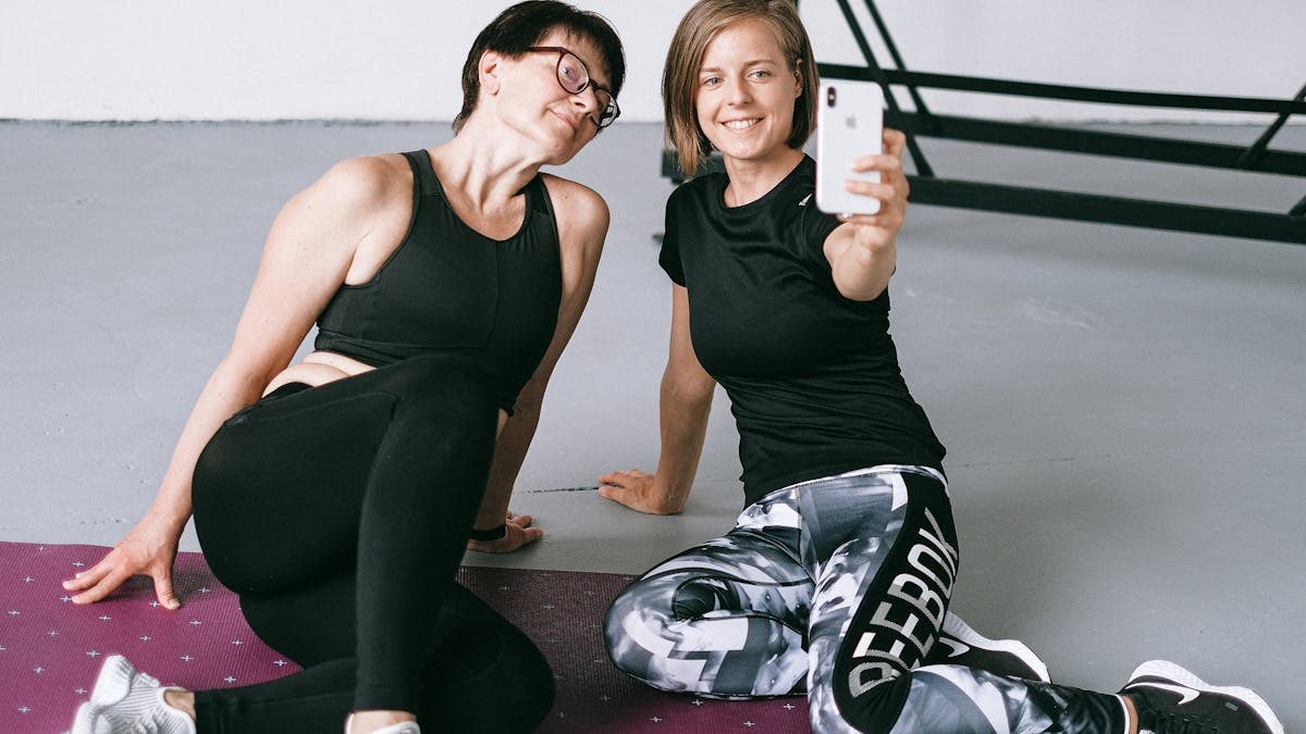 Two women in athletic clothing sitting on a yoga mat indoors, capturing a selfie post workout.